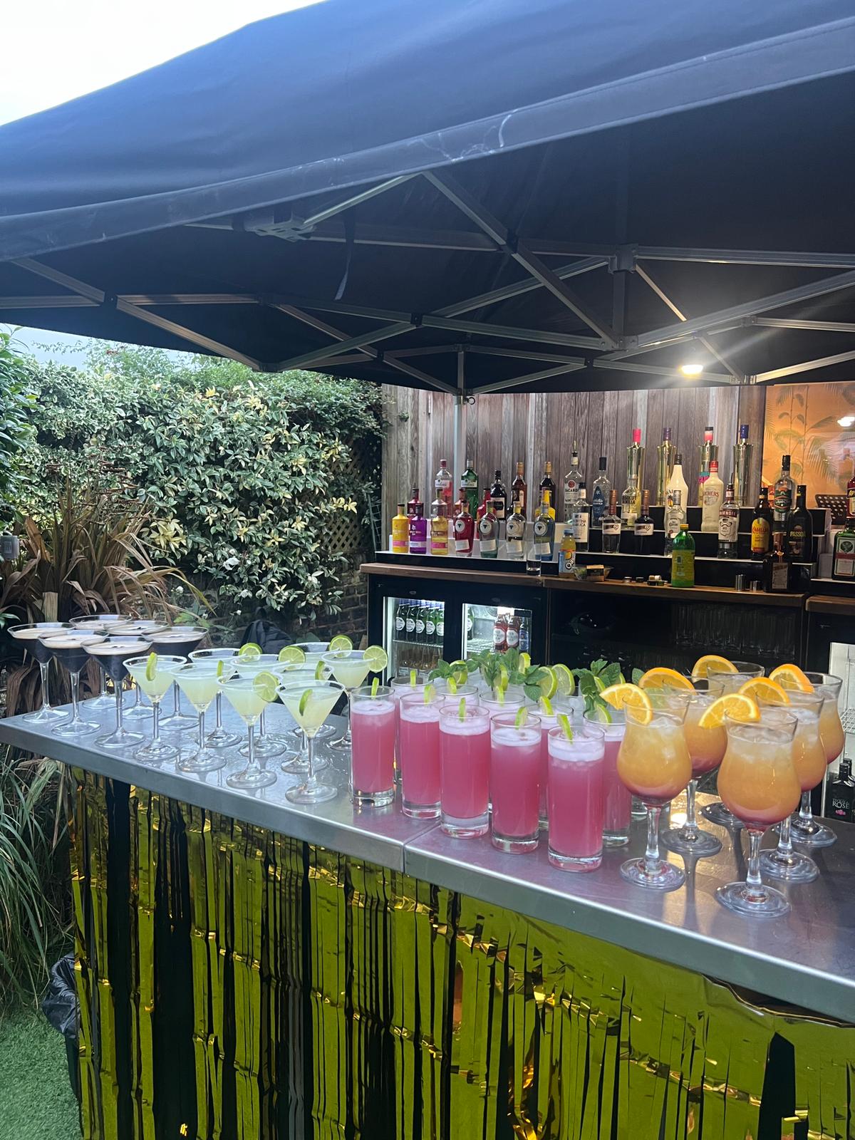 Rainbow of cocktails on a mobile bar with gold fringe at a garden party