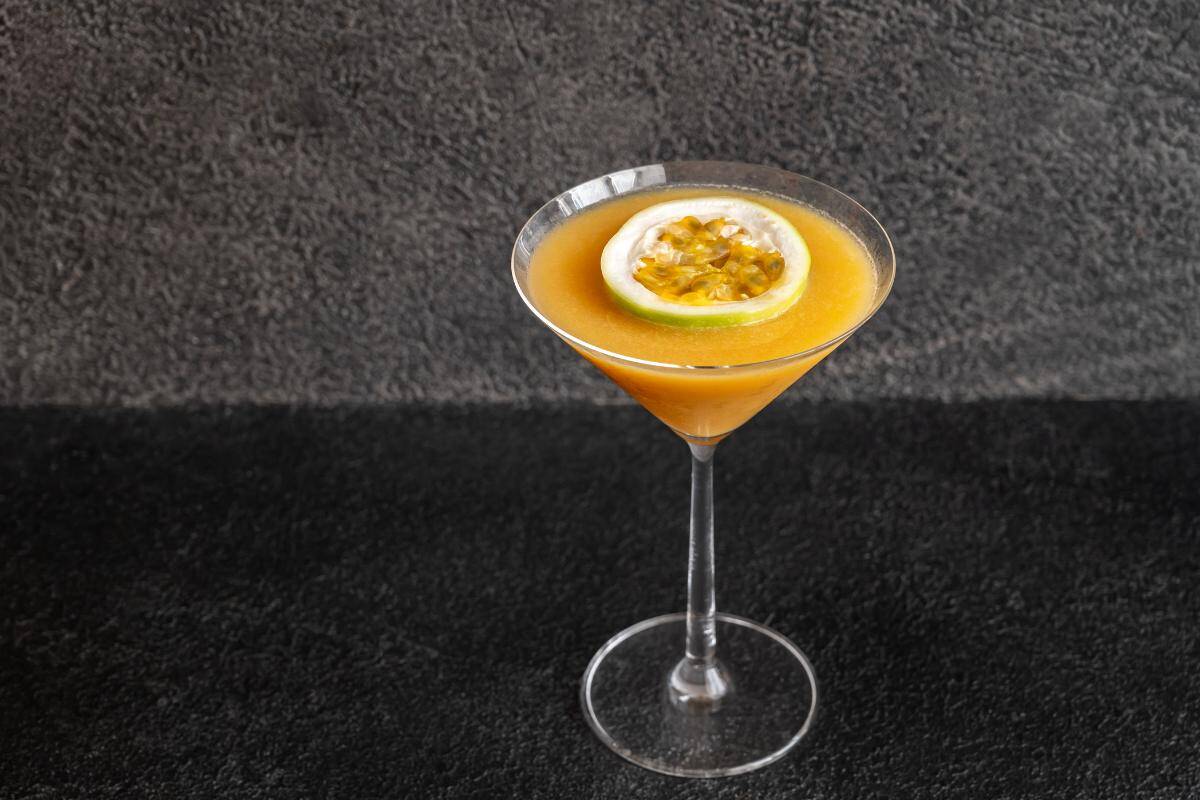 Passion Fruit Martini cocktail served in a classic martini glass, featuring vibrant orange colour and garnished with passion fruit and a slice of lemon on top.