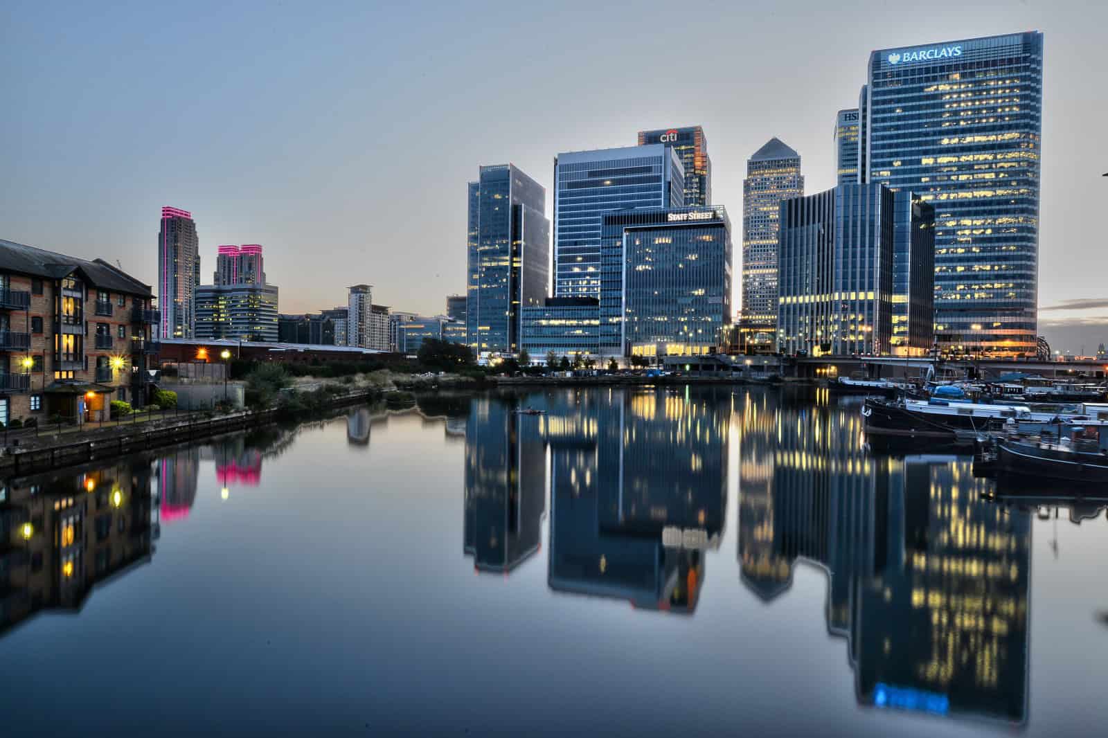 Modern London city skyline with illuminated skyscrapers reflected in the River Thames at dusk.