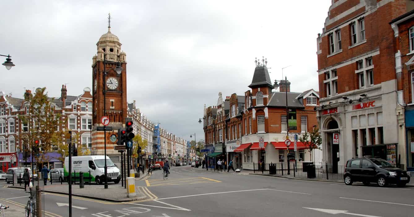Vintage red brick cityscape featuring historic clock tower and Victorian architecture in London, with busy street, pedestrians, and modern establishments.