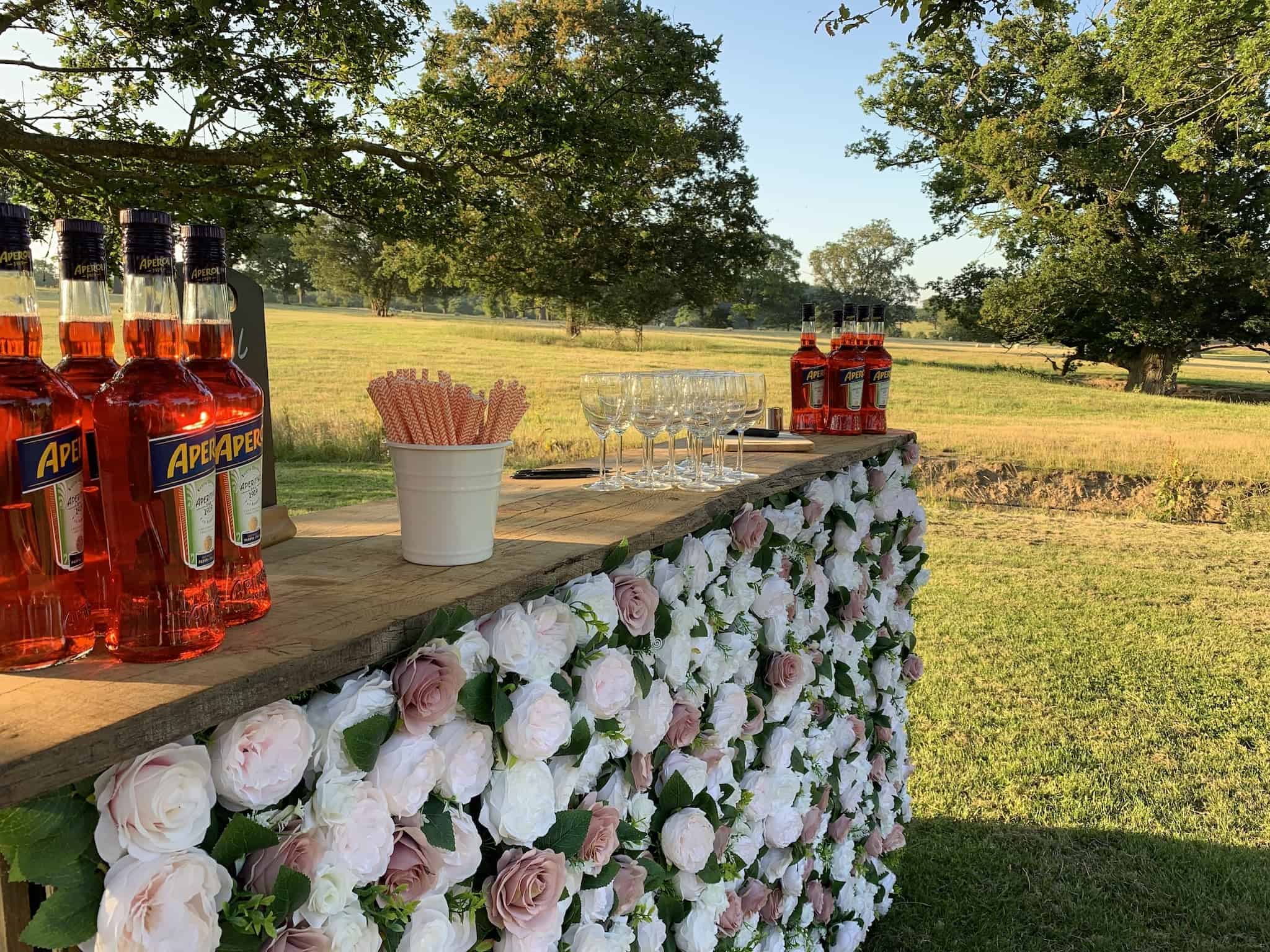 Bottles of Aperol spritz and cocktail glasses on a rustic outdoor bar decorated with white roses, set against a scenic green landscape on a sunny day.