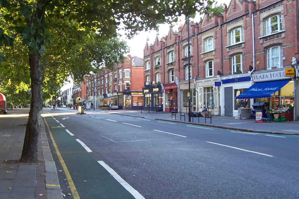 Modern street view of shops and cafes on a quiet London road during daytime, featuring brick buildings, storefronts, and trees, promoting The Sesh Bars mobile bar hire services in London and Berkshire.