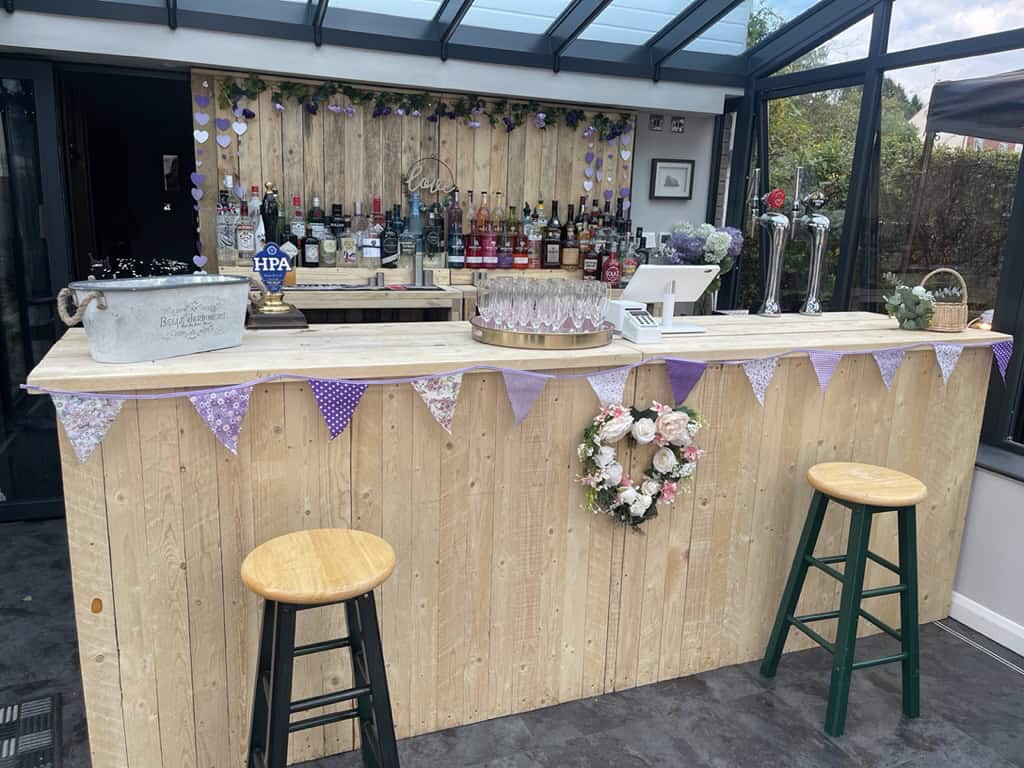 Vibrant outdoor wooden bar setup at The Sesh Bars Mobile Bar Hire London & Berkshire with floral decorations and a variety of alcohol bottles for events.