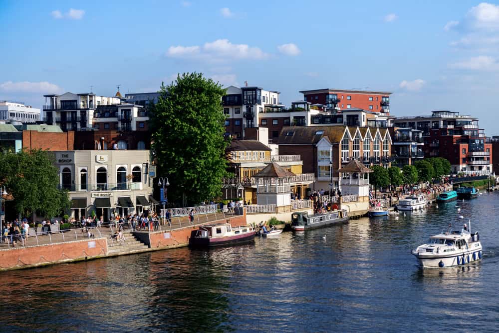 Boats moored along the riverside with modern buildings and greenery in the background in London, showcasing a lively riverside scene on a sunny day.