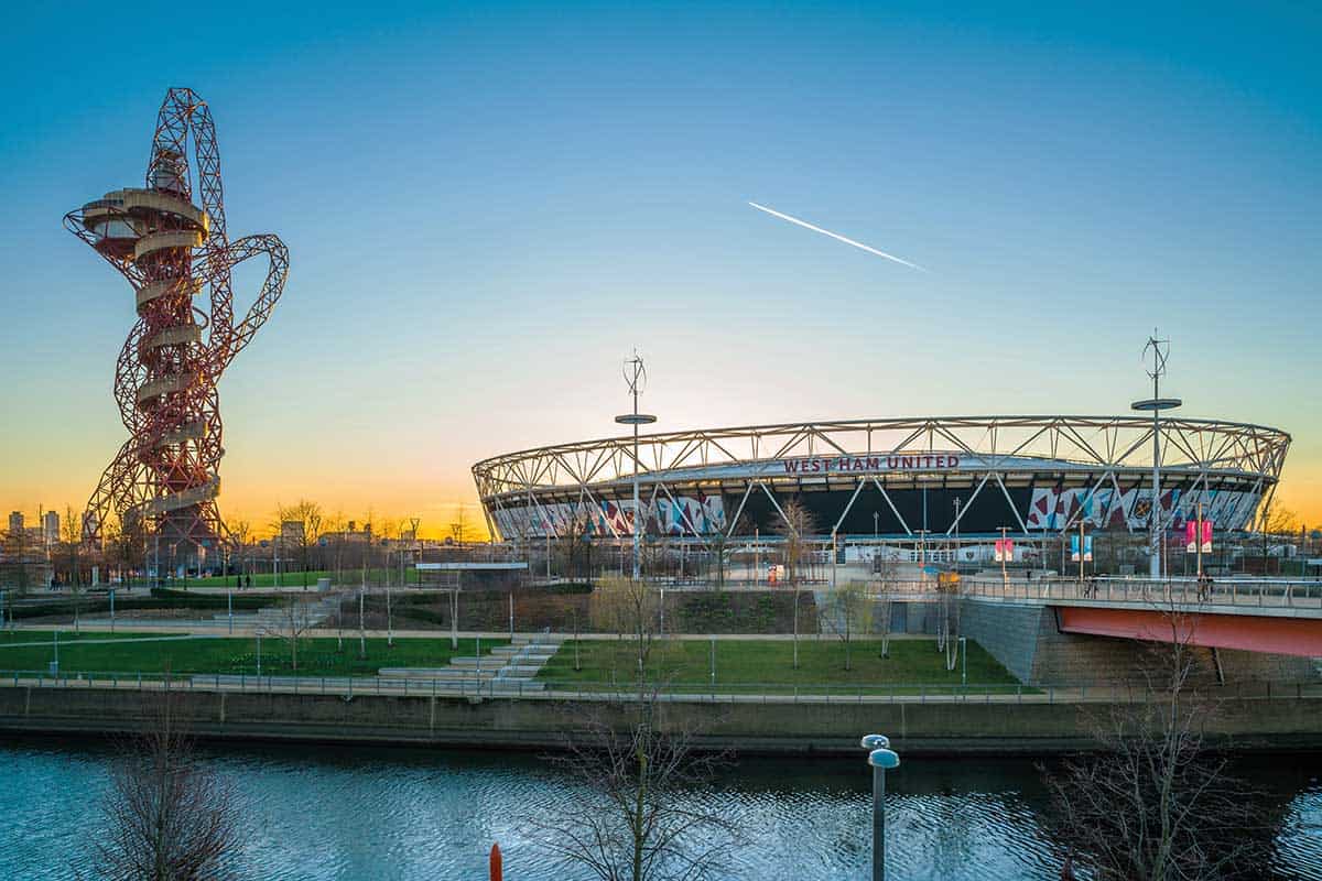 Modern London stadium and iconic structure with a waterfront view at sunset, showcasing vibrant sky colours behind The Sesh Bars mobile bar hire service.