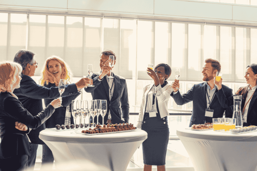 Prosecco toast at networking event with colleagues, standing around high tables with drinks and snacks in modern venue with large windows.