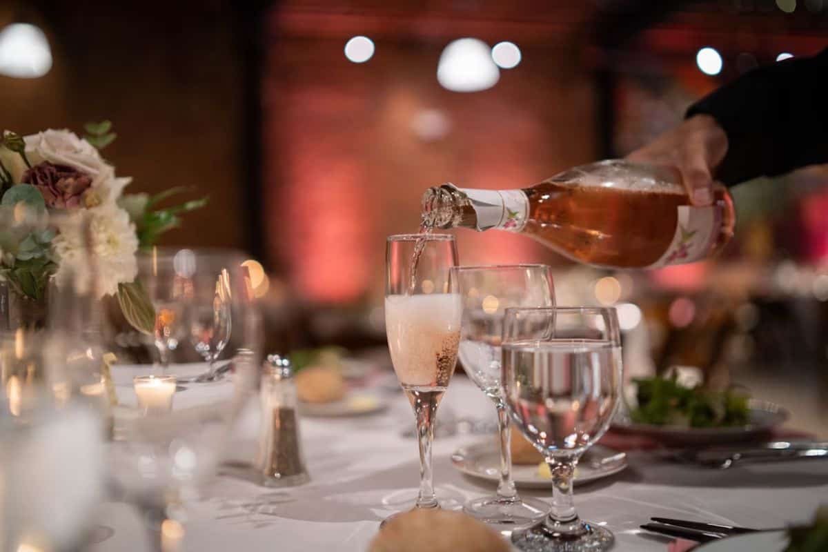A person pouring rosé wine into a glass at an elegant event with floral centrepieces and candlelit tables.
