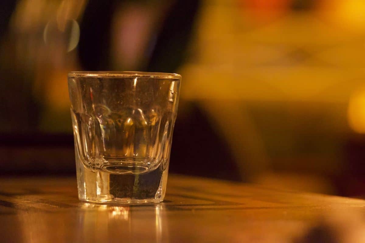A shot glass filled with a clear spirit, set on a wooden bar counter with warm ambient lighting in the background.